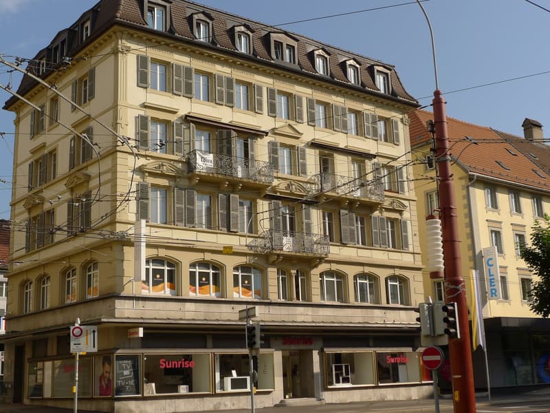 multi-story building, yellow facade, multiple windows, balconies on the second and third floor, tram wires above
