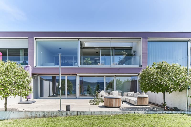 Two-story modern house with large glass windows and sliding doors, surrounded by greenery and with a balcony or terrace overlooking a scenic view. The exterior features a purple-colored facade with white trim. There is a patio area with comfortable outdoo