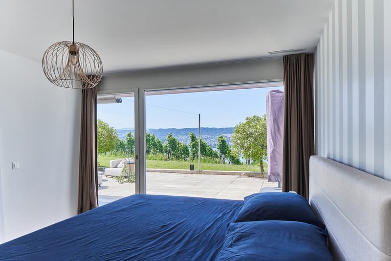 Spacious bedroom with large windows overlooking a scenic outdoor view of greenery and hills. The room features a blue bedspread, a hanging light fixture, and curtains on the sides of the windows.