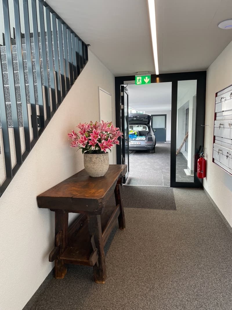 Car parked in garage with open car trunk, vase of flowers on a table in the hallway, steel railing on stairs