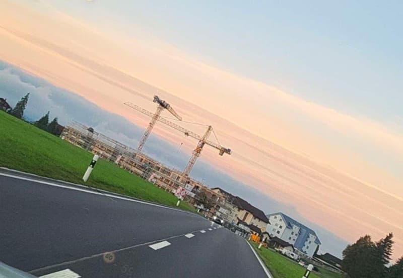 Construction site with cranes, road, green field, buildings in the background, sunset sky