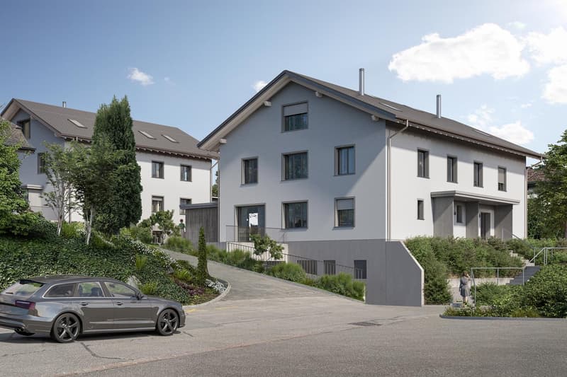 Multi-story apartment building with gray exterior, slanted roof, and several windows. There is a parking area in front of the building with a car parked. The building is surrounded by landscaping with trees and shrubs.