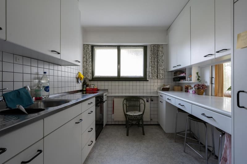 Modern kitchen with white cabinets, countertop, tile flooring, sink, oven, small table, chair, window, towels