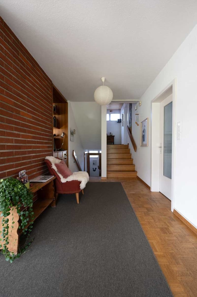 Carpeted floor, brick wall, wooden stairs, sofa with pillows, side table with plant, wooden cupboard, white ceiling