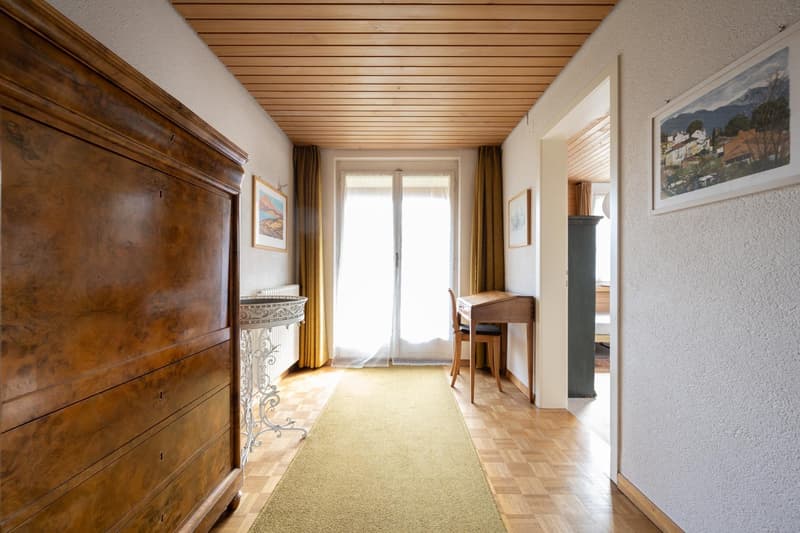 wooden ceiling, wooden floor, wooden dresser, yellow rug, white walls, large glass window, desk and chair, radiator, paintings on wall