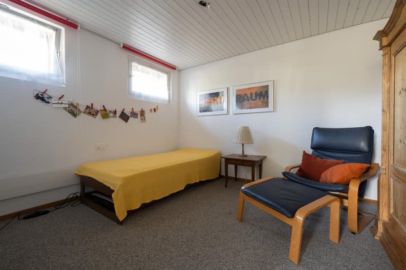 A small bedroom with a bed, bedside table, lamp, wall art, a wooden chair, and a cabinet