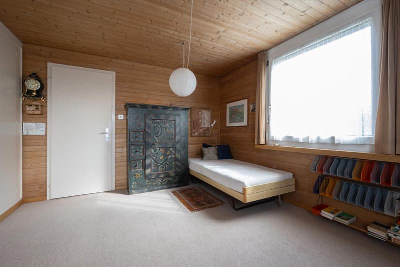 room with wooden walls, a white door, a white single bed, a rug, a wooden cabinet with drawers, and a shelf with books and baskets