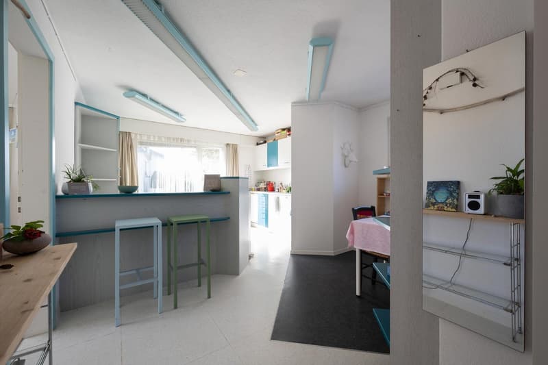 open plan kitchen with seating area, blue countertop, wooden bar stools, dining table, pink tablecloth, shelves, plant on the wall, white ceiling with blue lights