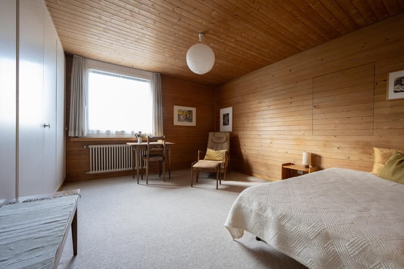 bedroom with white bed, wooden walls, wooden desk, wooden chair, radiator, picture frames, table lamp, carpet, bench