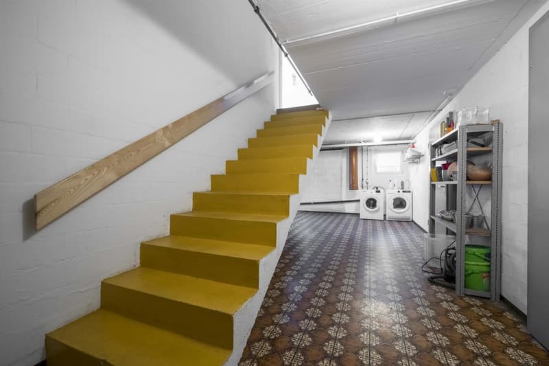Yellow painted staircase, white walls, tiled floor, utility area with white front-load washers, metal storage shelving, green buckets, glass containers, and cleaning supplies.