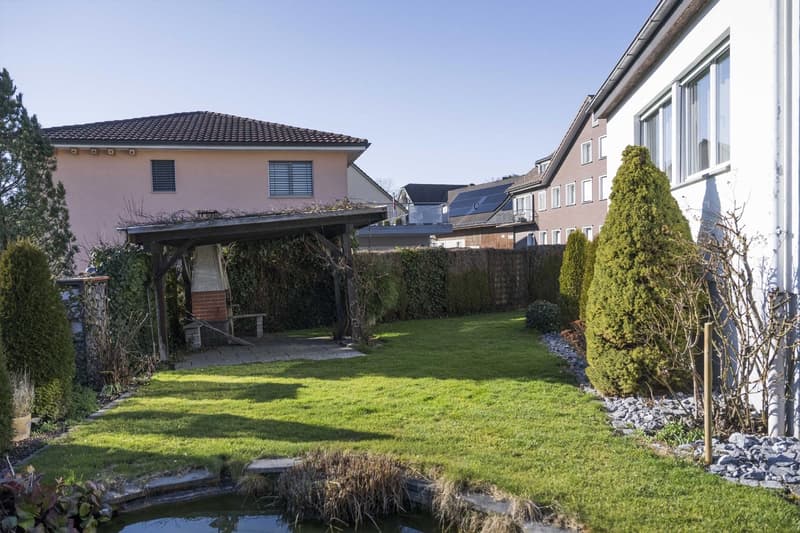 Large open backyard with green grass, small pond, outdoor seating under gazebo, brick seating, wooden bench, and well-maintained shrubs and plants.