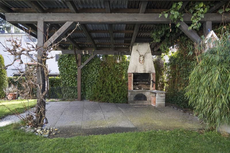 wooden pergola covering brick fireplace with overhanging roof, stone patio, shrubs, green grass