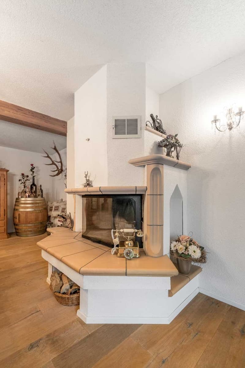 Living room with white walls and wooden floors, featuring a corner fireplace with a mantle, wooden telephone, and decorative items such as flowers and antlers
