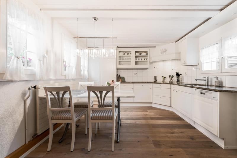 All-white cabinets and countertops, wooden floor, chandelier, dining table with chairs, open shelving, windows with white curtains, countertop with kitchen sink and faucet.