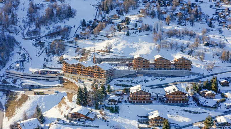 Several modern chalets, some under construction, set in a snowy alpine landscape with roads and a ski lift in the background.