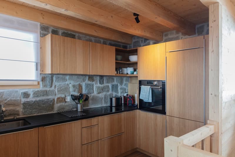 modern kitchen with wooden cabinets, black countertops, stainless steel sink, built-in oven, wooden beams, stone wall