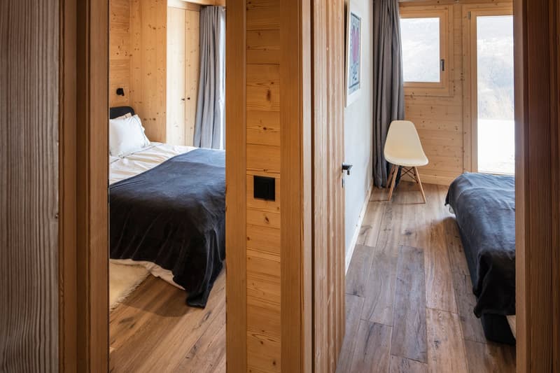 bedroom with wooden walls, two beds, white chair, large windows, wooden floor