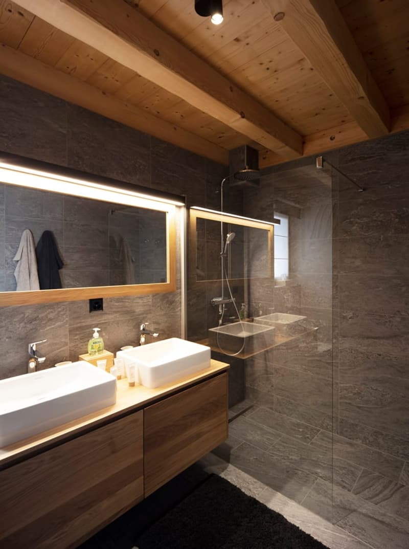 modern bathroom with wooden ceiling, gray tiles, wooden vanity, two sinks, mirror, shower, black rug