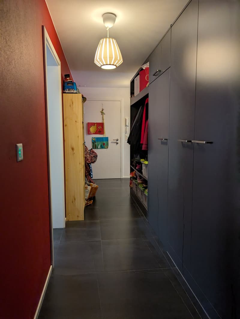 Wide hallway with red wall, cabinets, storage boxes, hanging decorations, and an entrance to another room