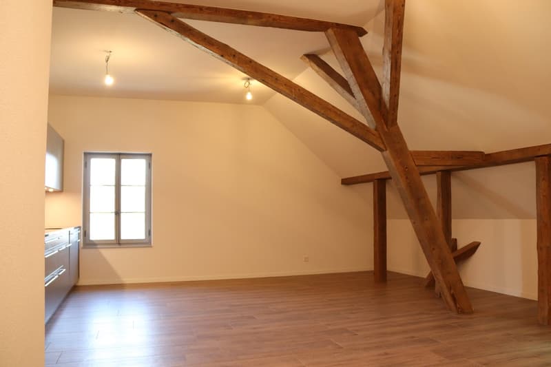 Empty attic room with wooden beams, hardwood floors, a window, and a kitchen area.