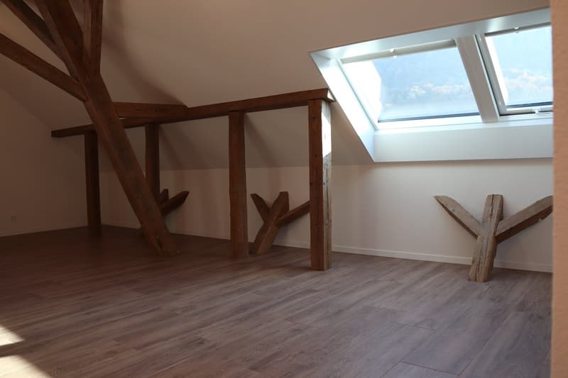 Empty room with wooden beams and a slanted ceiling with windows.