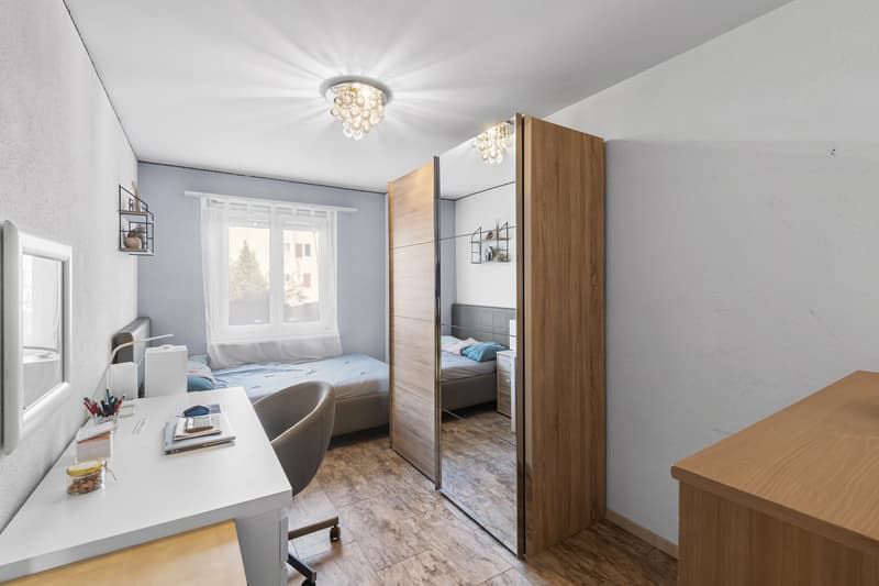 a desk with a chair, a mirror on the wall, a bed with a mattress and pillows, a bedside cabinet, a shelf on the wall, and a chandelier