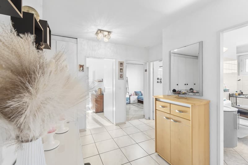 A hallway leading to a bedroom with a bed, a cabinet, a mirror, a bathroom, and a white tiled floor.