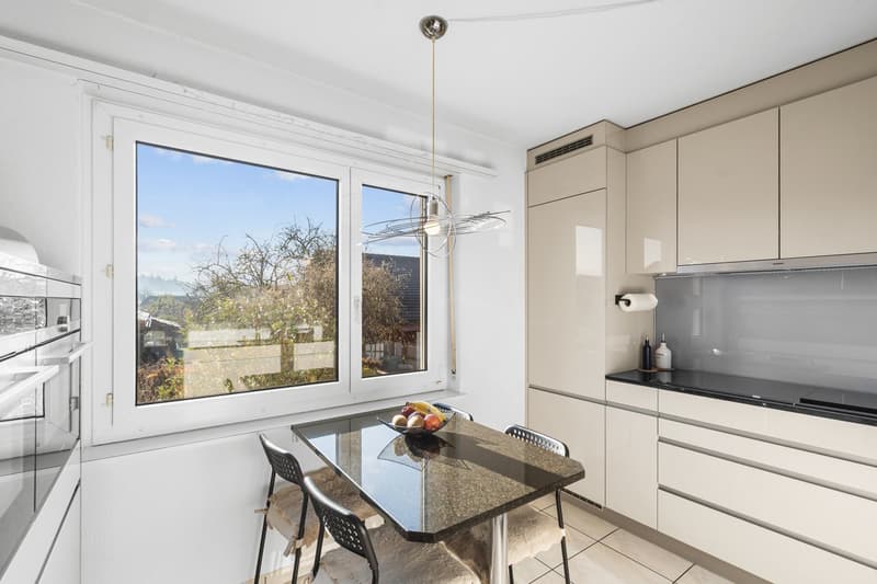 White walls, black tiled floor, large window with view, glass table, hanging lamp, white kitchen