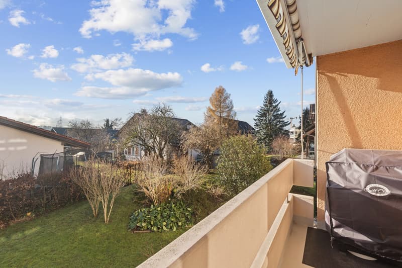 An outdoor balcony or terrace with a railing and a covered area. There is a grill cover nearby, and a lush green lawn with plants and trees visible in the background. The sky is clear with a few clouds.