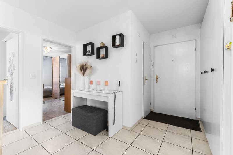 Entrance hall with a white table, black stool, framed pictures on wall, tile flooring, white doors, black doormat.