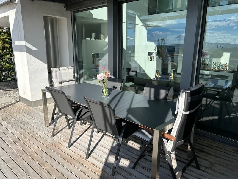 A modern outdoor dining area with a black table and chairs on a wooden deck, enclosed by large glass walls and doors, with a view of the outdoors.