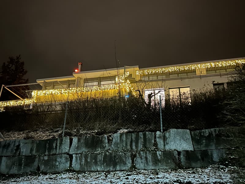 A building with a balcony or terrace decorated with string lights, set against a dark night sky with a stone wall in the foreground.