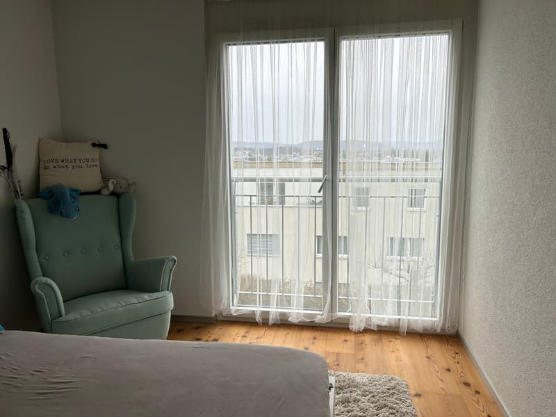 Bedroom with large windows, white curtains, green armchair, wooden floor, and a decorative pillow on the bed