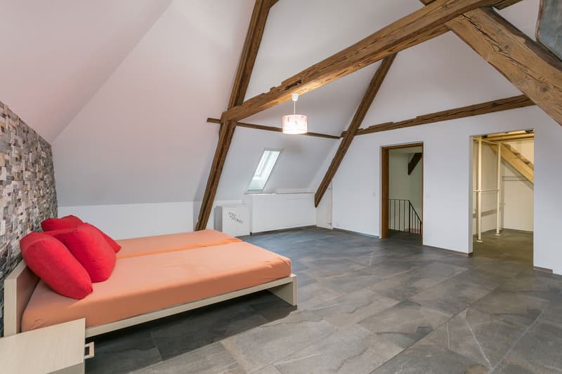 Spacious attic bedroom with exposed wooden beams, gray tiled flooring, a large window, and a red and orange bedding set on a platform bed. There is also a small closet or storage space visible in the room.