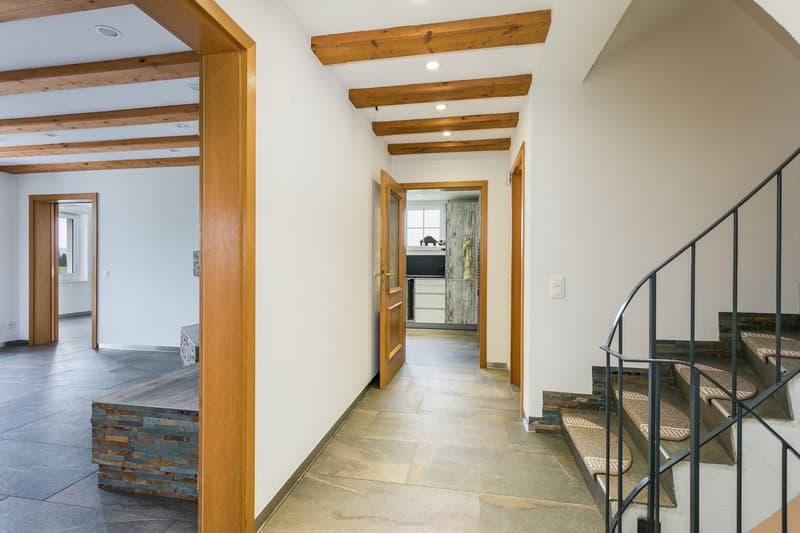 The image shows a spacious interior with a hallway featuring exposed wooden beams on the ceiling, a stone fireplace, and a staircase with a metal railing. The floors are tiled, and there are wooden doors leading to other rooms. The overall style appears t