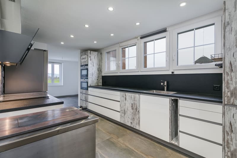 Modern kitchen with white cabinets, black countertops, stainless steel appliances including a dishwasher, large windows providing natural light, and a tiled floor.
