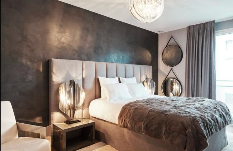 Dark wall with metal texture, bed with beige cover, nightstand with modern lamp, two round mirrors, large white window with curtain, white armchair