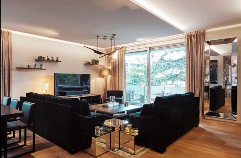 Living room with black sofas, glass coffee table, dining table, television, modern lighting fixtures, balcony visible through the sliding doors, large windows, wooden flooring, beige walls