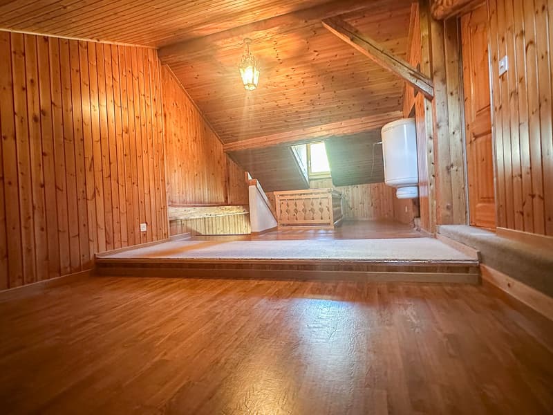 A wooden attic room with a sloped ceiling, carpeted floor, wooden walls, ceiling lamp, wooden storage box, wall-mounted water heater, and an attic window.