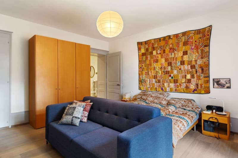 Blue sofa, wooden cabinet, bed, nightstand, lamp, decorative throw blanket, open door, tiled floor, quilted tapestry on wall