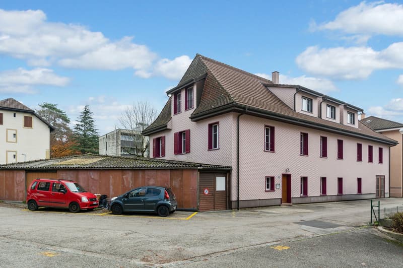 2 story house, pink facade, brown roof, large windows, brick attached garage, multiple parking spots, red and grey cars parked.