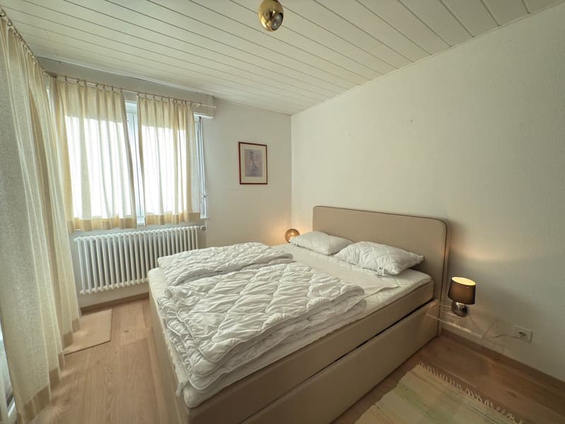 double bed, two pillows, a nightstand with a lamp, white linen, wooden floor, white walls, white curtains, a window with a radiator