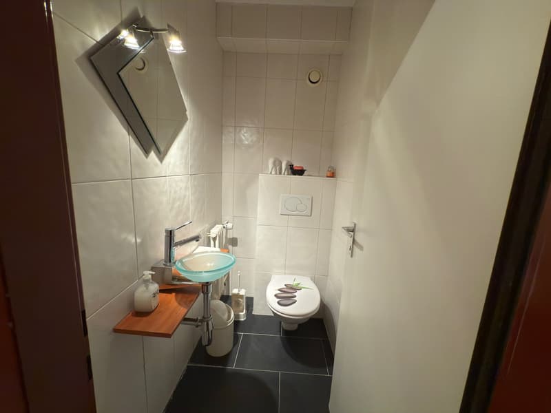 Compact bathroom with a sink, toilet, wooden shelf, and white tiled walls and floors