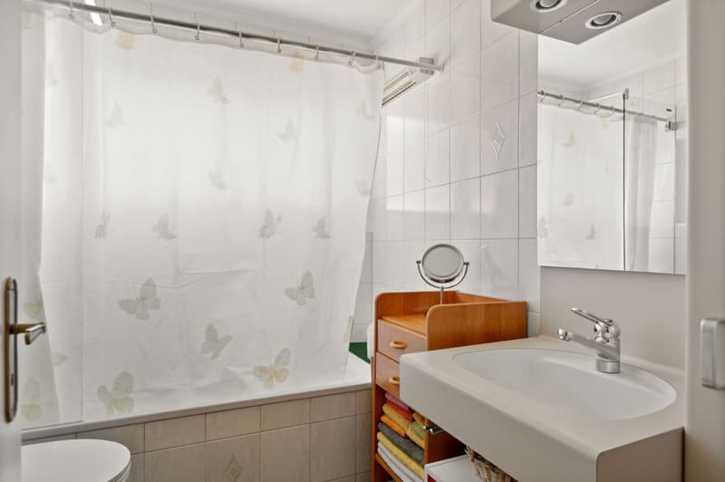 White tiled walls, bathtub with shower curtain, vanity with sink, mirror, wooden cabinet, white towels