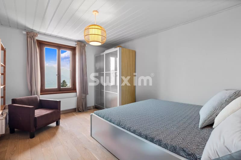 Chambre lumineuse avec lit double, fauteuil marron, et grande fenêtre offrant une vue dégagée.