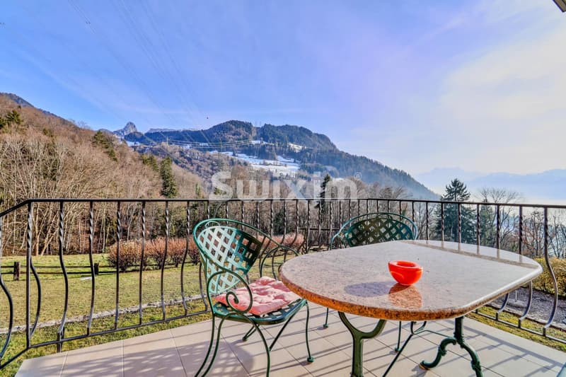 Une terrasse avec une table et deux chaises en fer forgé, surplombant un paysage montagneux et des arbres.
