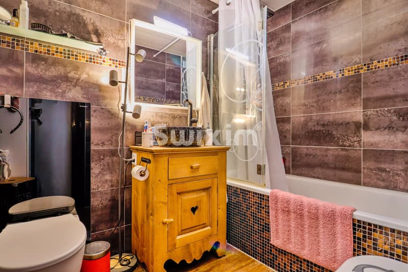 Une salle de bain chaleureuse avec des carreaux marron, une baignoire, un lavabo sur meuble en bois et une serviette rose accrochée.