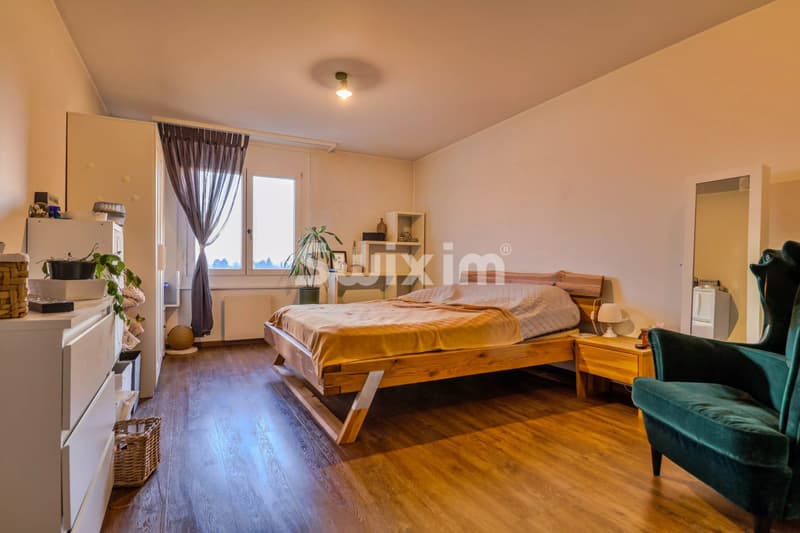 Une chambre spacieuse avec un lit en bois, des meubles modernes, un fauteuil vert et une grande fenêtre laissant entrer la lumière naturelle.