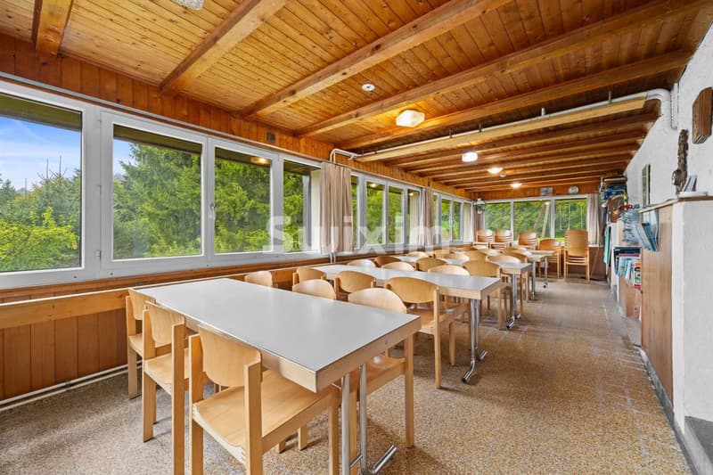 Une salle à manger lumineuse avec de grandes fenêtres, des tables en bois et des chaises, donnant sur un paysage verdoyant.