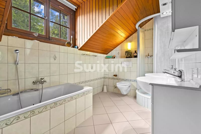 Une salle de bain avec un toit en pente, une baignoire, un lavabo et une fenêtre en bois offrant une vue sur l'extérieur.